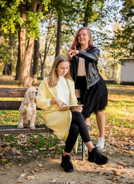 Two cheerful female best friends in casual wear using tablet in autumn sunny park Happy teen girls outdoors