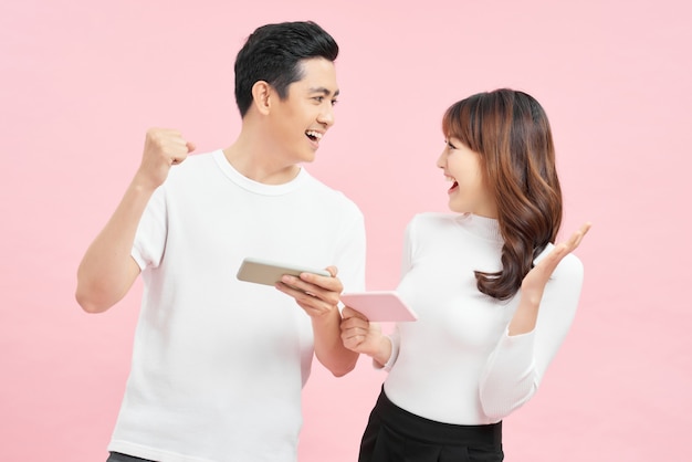 Two cheerful friends standing isolated over pink background, playing games on mobile phone.