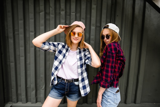 Two cheerful funny girl wearing checkered shirts posing against street wall at the street.