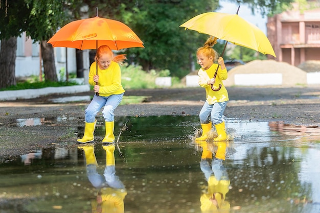 Two cheerful girls with yellow umbrellas run through the\
puddles in rubber boots