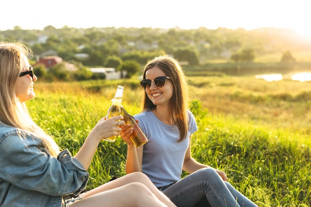 Two cheerful girls and young friends with sunglasses, drinking beer and enjoying the time spent together at sunset.