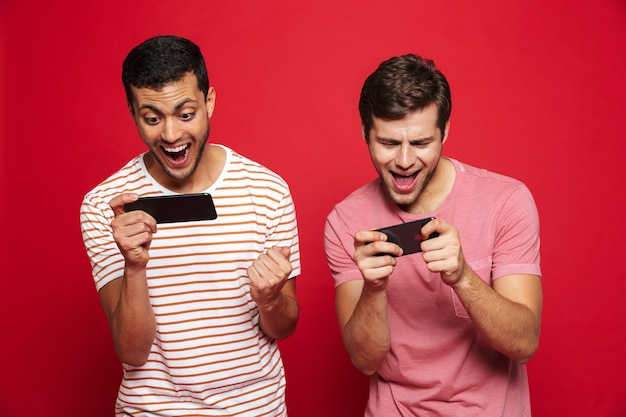Photo two cheerful men friends standing isolated over red wall, playing games on mobile phones