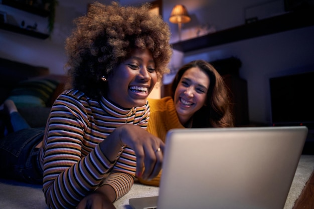 Foto due donne multietniche allegre che guardano il computer sdraiate sul tappeto nel salotto buio