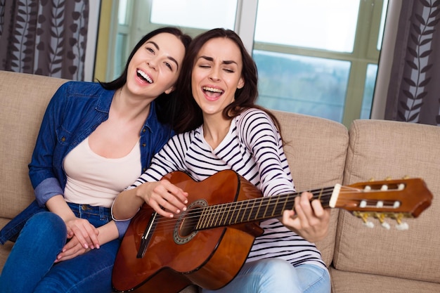 Two cheerful and smiling girls in ordinary clothes sit at home on the couch and sing their favorite songs to the guitar