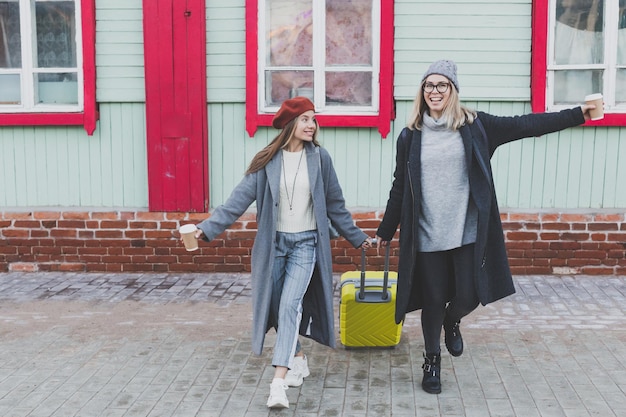 秋または春のティムの街の通りを笑顔でスーツケースを持って歩く2人の陽気な観光客の女性