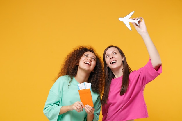 Two cheerful women friends european african girls in pink green clothes posing isolated on yellow background. people lifestyle concept. mock up copy space. hold passport boarding pass ticket airplane