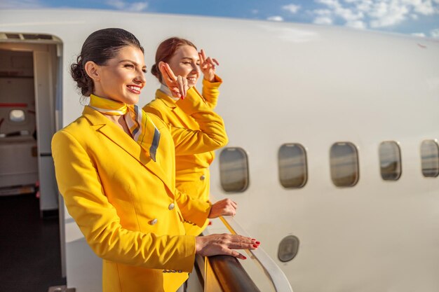 Two cheerful women stewardesses in aviation uniform doing salute gesture and smiling while standing near aircraft door