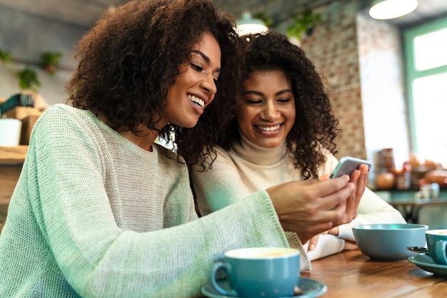 Two cheerful young african girlfriends sitting at the cafe drinking coffee, looking at mobile phone