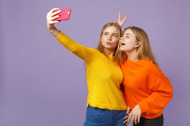 Two cheerful young blonde twins sisters girls in colorful clothes doing selfie shot on mobile phone isolated on pastel violet blue wall . People family lifestyle concept. 