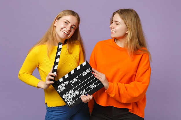 Two cheerful young blonde twins sisters girls in colorful clothes hold classic black film making clapperboard isolated on violet blue wall. People family lifestyle concept.