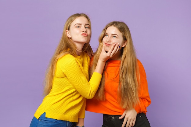 Two cheerful young blonde twins sisters girls in vivid colorful clothes having fun, fooling around isolated on pastel violet blue wall . people family lifestyle concept