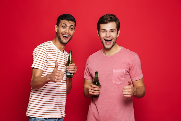 Two cheerful young men standing isolated over red wall, drinking soda water from bottles, thumbs up
