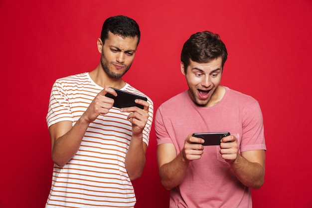 Two cheerful young men standing isolated over red wall, playing games on mobile phone