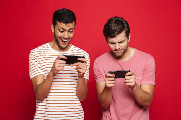 Two cheerful young men standing isolated over red wall, playing games on mobile phone