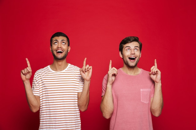 Two cheerful young men standing isolated over red wall, pointing at copy space