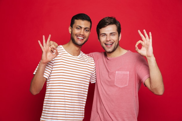 Two cheerful young men standing isolated over red wall, showing ok