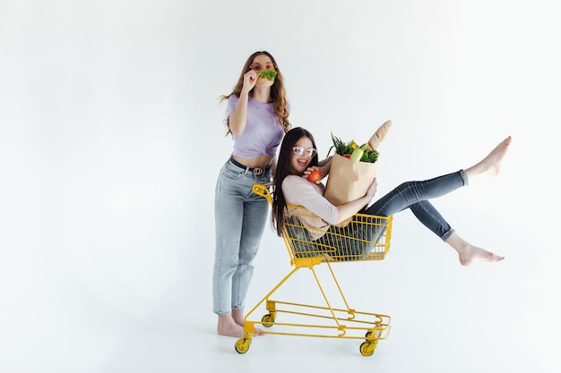 Two cheerful young women in colorful trendy outfits smiling and having fun with trolley