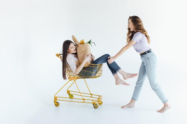 Two cheerful young women in colorful trendy outfits smiling and having fun with trolley