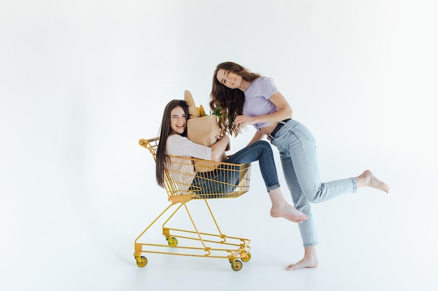 Two cheerful young women in colorful trendy outfits smiling and having fun with trolley