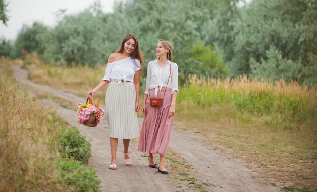 Две веселые молодые женщины в одежде в стиле ретро идут по площадке с корзиной для пикника и разговаривают.