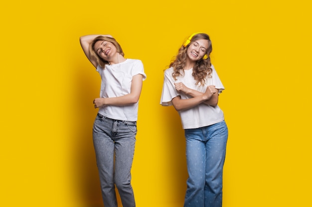 Two cheering sisters are listening to music using headphones and dancing on a yellow wall