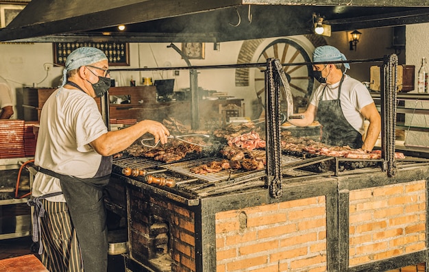 Two chefs wearing face masks and hair nets cooking meat in the grill and using meat tongs.