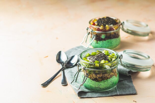 Two chia seed pudding with matcha green tea kiwi and granola orange in glasses on light pink concrete background healthy breakfast square image