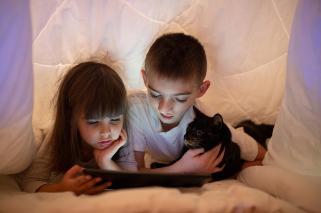 Two children boy and girl, brother and sister, friends lying under a white blanket on the bed reading playing studying on a tablet computer phone with black cat