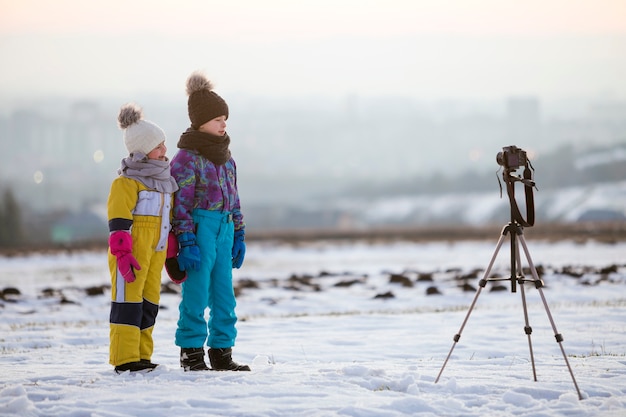 Due bambini ragazzo e ragazza divertirsi fuori in inverno giocando con la macchina fotografica su un treppiede sul campo coperto di neve.