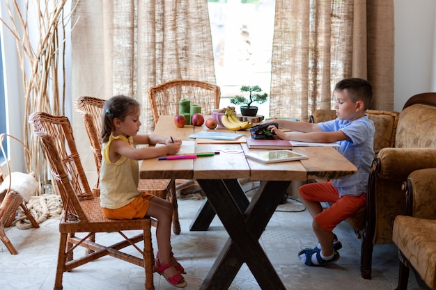 Two children, a boy and a girl, do their homework, sitting at a large wooden table in the dining room of their house, they draw and write, they develop their creativity.