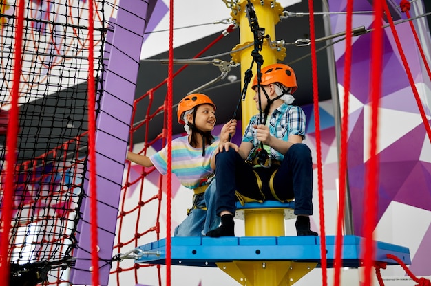 Two children in helmets climb on tightropes in entertainment\
center. boy and girl having fun on ropes in climbing area, kids\
spend the weekend on playground, happy childhood