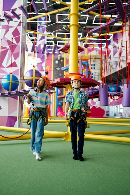 Two children in helmets climb on zip line in entertainment\
center, young climbers, front view. boy and girl having fun on\
ropes in climbing area, kids spend the weekend on playground, happy\
childhood