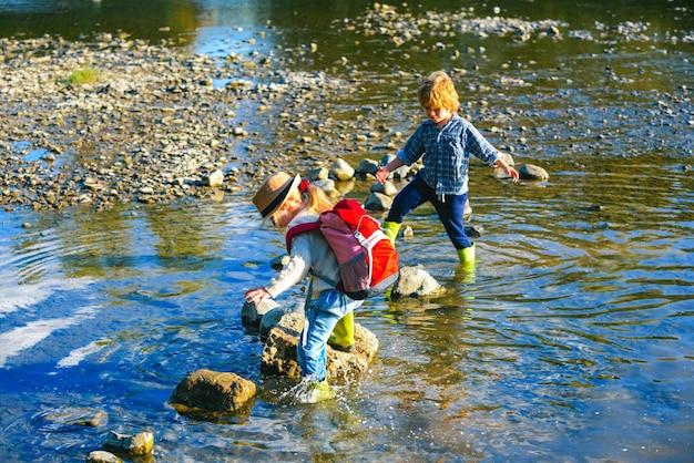 Two children with a backpack stands near a mountain river Boy and girl looks at the creek The kids w