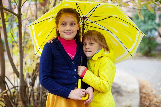 Two children with yellow umbrella stand side by side on street on autumn dayxDxA