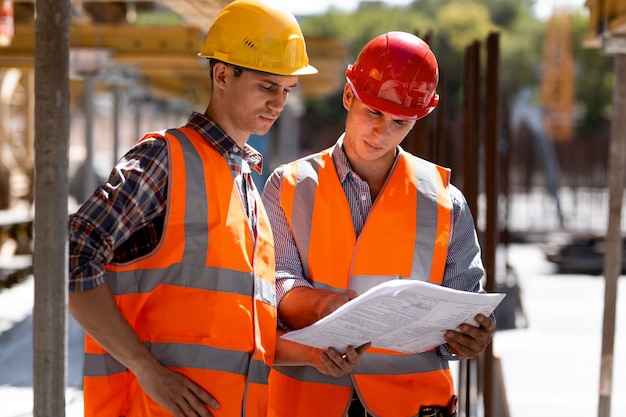 Two civil engineers dressed in orange work vests and helmets explore construction documentation on the building site near the wooden building constructions