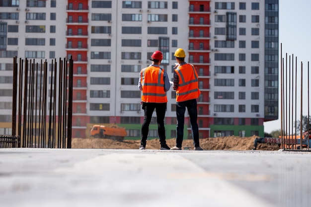 Two civil engineers dressed in orange work vests and helmets talk about the construction process on the building site against the background of a multi-storey building .