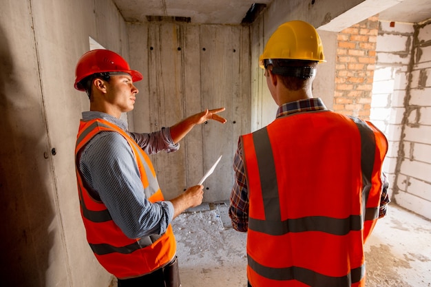 Two civil engineers dressed in orange work vests and helmets walk inside the building under construction .