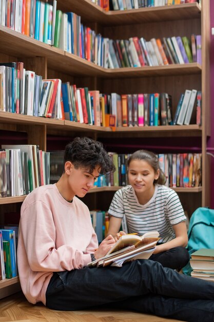 Two clever teenage students sitting on the floor of college library by bookshelf and discussing passage from story