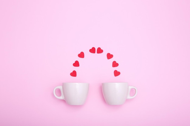 Две кофейные чашки и красные деревянные сердца на розовом фоне. Концепция романтического подарка или предложения руки и сердца.