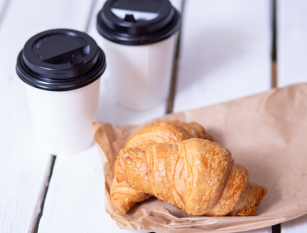Two coffees in paper cups and croissants on a white wooden table