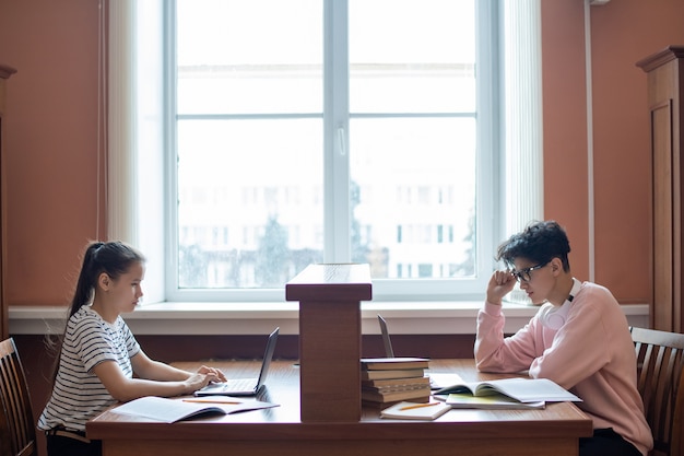 Two college learners sitting by desks in front of one another and looking at laptop displays while surfing in the net