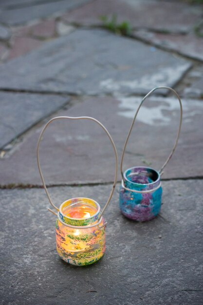 Two colorful glass jars with wire handle candle lamps on stone outdoor tile, kids activities and handmade idea concept vertical