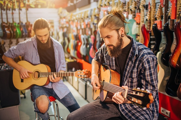 Two concentrated young man playing on acoustic guitars. They sit on stools in room full on electric guitars. Bearded guys play together.