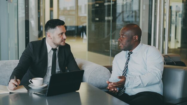 Two confident businessmen discussing about partnership during meeting in modern glassy cafe