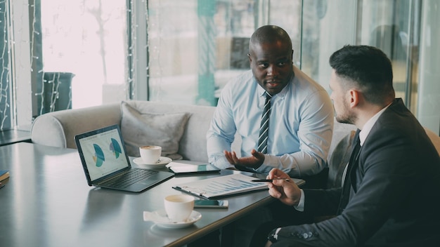 Two confident businessmen looking at diagrams and graphs on laptop computer and discussing the financial report and statistics of their business project in modern cafe during lunch time