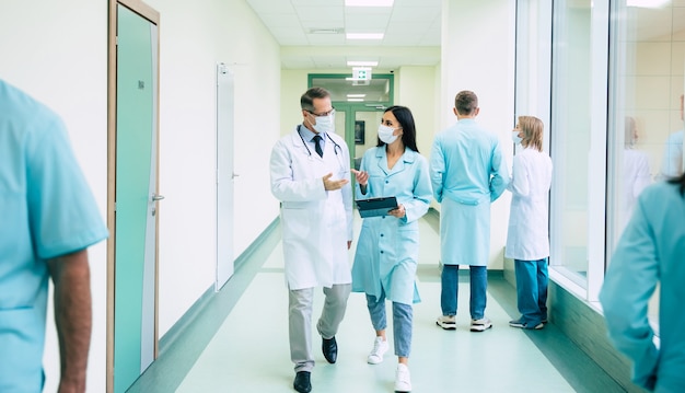 Two confident doctors in safety medical masks have a discussion about some treatment methods during walking the hospital corridor with other colleagues around them