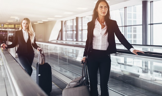 Two confident women wearing formal suits standing on moving walkway in airport