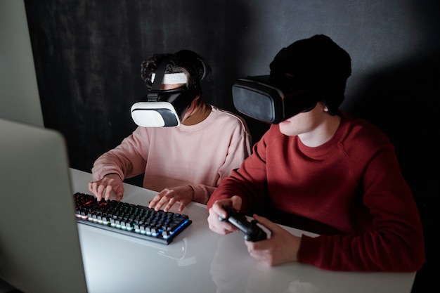 Двое современных подростков в гарнитурах vr сидят за столом перед экраном компьютера, играя в игры в темноте