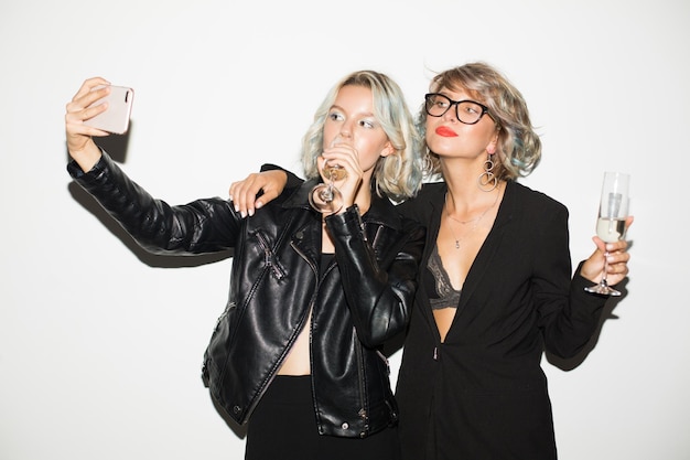 Two cool girls in black jackets with glasses of champagne in hands dreamily taking photos on cellphone over white background isolated