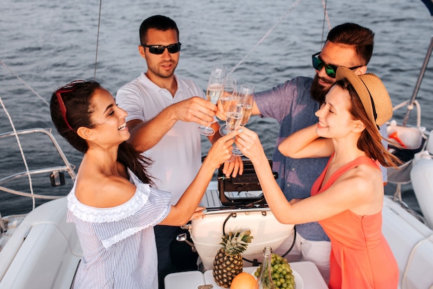 Two couples stand in front of each other and cheers with glasses of champaigne. They look at it and smile. People sailing on yacht.